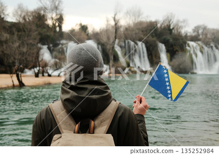 Traveler seen from behind holding Bosnia and Herzegovina flag while facing Kravica Waterfall. Exploration and reflection concept with wild nature, water scenery, and winter calm Traveler seen from behind holding Bosnia and Herzegovina flag while facing Kravica Waterfall. Exploration and reflection concept with wild nature, water scenery, and winter calm 135252984