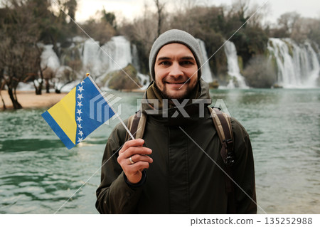 Smiling man holding Bosnia and Herzegovina flag in front of Kravica Waterfall. Travel portrait with national identity, natural landscape, winter mood, and calm outdoor atmosphere Smiling man holding Bosnia and Herzegovina flag in front of Kravica Waterfall. Travel portrait with national identity, natural landscape, winter mood, and calm outdoor atmosphere 135252988