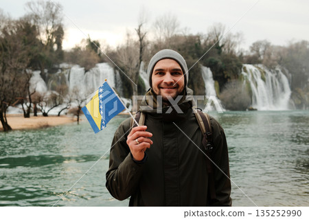 Smiling man holding Bosnia and Herzegovina flag in front of Kravica Waterfall. Travel portrait with national identity, natural landscape, winter mood, and calm outdoor atmosphere Smiling man holding Bosnia and Herzegovina flag in front of Kravica Waterfall. Travel portrait with national identity, natural landscape, winter mood, and calm outdoor atmosphere 135252990