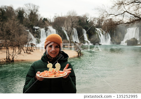 Smiling woman holding a cake in the form of sandwiches with red fish caviar with candles in front of Waterfall. Travel celebration concept with nature landscape, winter atmosphere, and joyful mood 135252995