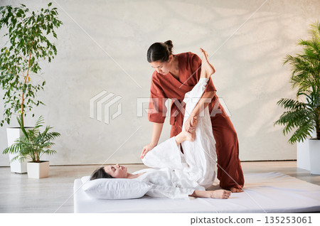 Masseuse making thai yoga massage. Woman therapist doing traditional massage treatment, stretching female patient's body. Client lying on mat, enjoying hand massage in calm atmosphere. 135253061