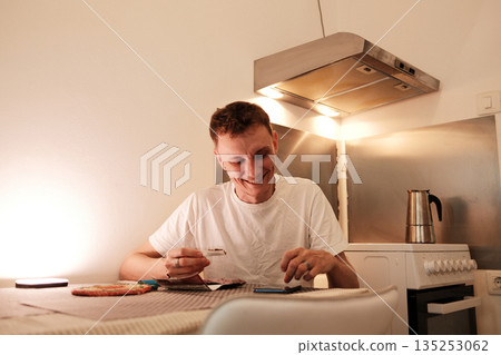 A man smiles while rolling a hand rolled cigarette at the kitchen table. The concept shows a relaxed moment of personal routine and simple enjoyment 135253062