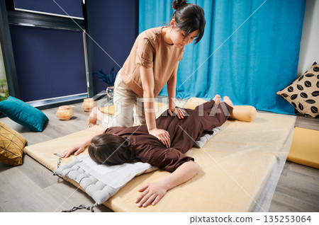 Masseuse making thai yoga massage. Woman therapist doing traditional massage treatment for female patient indoors. Client lying on mat, meditating, enjoying hand massage in calm atmosphere. Masseuse making thai yoga massage. Woman therapist doing traditional massage treatment for female patient indoors. Client lying on mat, meditating, enjoying hand massage in calm atmosphere. 135253064