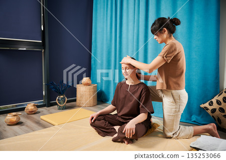 Masseuse making thai yoga massage. Woman therapist doing traditional massage treatment for female patient indoors. Client sitting on mat, meditating, enjoying hand massage in calm atmosphere. 135253066