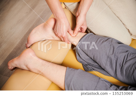 Masseuse making thai yoga massage. Woman therapist doing traditional massage treatment for male patient's foot. Close up. Client lying on mat, meditating, enjoying hand massage in calm atmosphere. 135253069