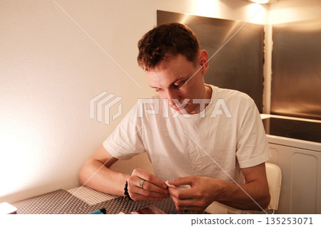 A man sits at the table and carefully prepares a hand rolled cigarette. The concept highlights a quiet personal routine and focused manual work 135253071