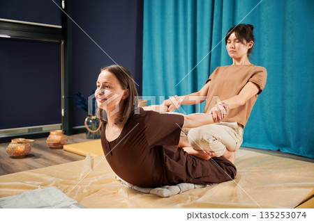 Masseuse making thai yoga massage. Woman therapist doing traditional massage treatment, stretching female patient's body. Client lying on mat, enjoying hand massage in calm atmosphere. 135253074