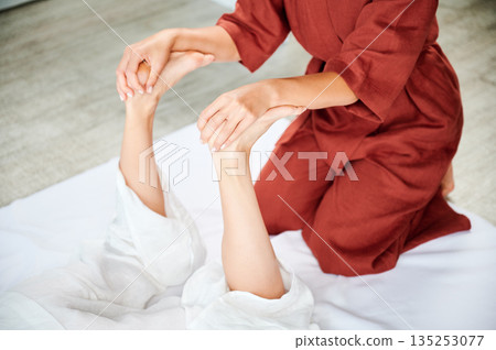 Masseuse making thai yoga massage. Woman therapist doing traditional massage treatment, stretching female patient's body. Client lying on mat, enjoying hand massage in calm atmosphere. Cropped view. 135253077