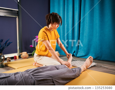 Masseuse making thai yoga massage. Woman therapist doing traditional massage treatment for male patient indoors. Client lying on mat, meditating, enjoying hand massage in calm atmosphere. 135253080