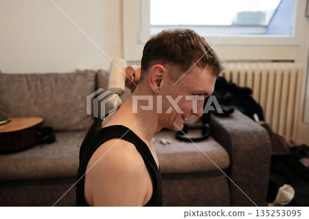 A man uses a massage gun on his neck while sitting on the floor. The scene conveys home relaxation concept 135253095