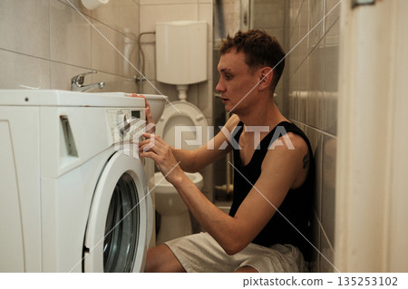 A man adjusts the settings on a washing machine while sitting on the bathroom floor. The scene represents practical home routine concept A man adjusts the settings on a washing machine while sitting on the bathroom floor. The scene represents practical home routine concept 135253102
