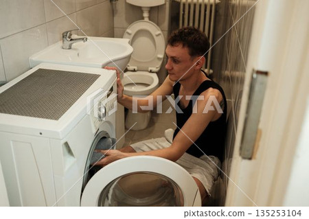 A man places a towel inside a washing machine in a small bathroom. The scene reflects everyday home routine concept A man places a towel inside a washing machine in a small bathroom. The scene reflects everyday home routine concept 135253104