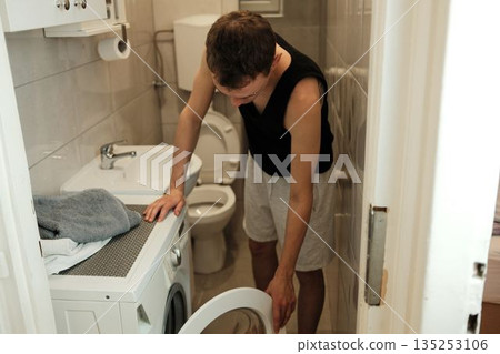 A man opens the door of a washing machine in a small bathroom. The moment shows simple home routine concept A man opens the door of a washing machine in a small bathroom. The moment shows simple home routine concept 135253106