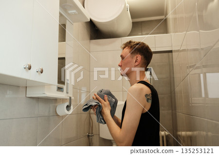 A man holds a towel while standing at the bathroom sink under soft light. The concept highlights personal hygiene and morning care. A man holds a towel while standing at the bathroom sink under soft light. The concept highlights personal hygiene and morning care. 135253121