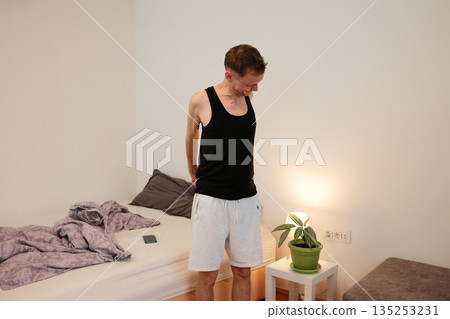 A man stands beside the bed stretching lightly after waking up. The concept shows a natural morning routine and physical awakening. 135253231