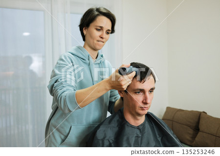 A woman uses a trimmer to shorten a man s hair during a home haircut. The moment reflects personal grooming and the concept of everyday care 135253246