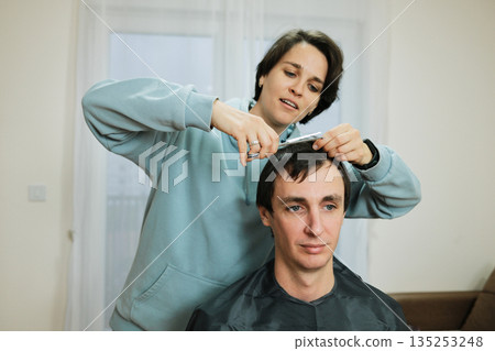 A woman trims a man s hair at home using scissors while he sits calmly. The scene presents everyday grooming and the concept of casual personal care A woman trims a man s hair at home using scissors while he sits calmly. The scene presents everyday grooming and the concept of casual personal care 135253248