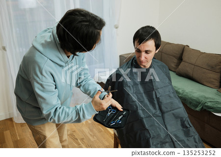 A man sits covered with a haircut cape while woman prepares scissors at home. The scene shows casual grooming and the concept of simple personal care 135253252