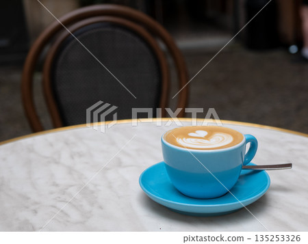 Heart-shaped latte art in a blue ceramic cup on a marble cafe table. A cozy outdoor coffee moment with elegant details and warm ambiance 135253326