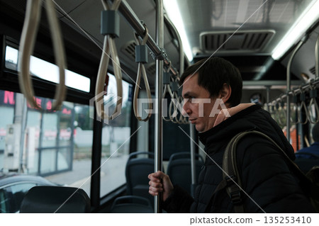 A man stands inside a bus and holds the vertical pole while riding. The scene represents public transport and the concept of routine movement 135253410