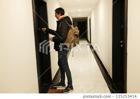A man stands in a hallway with a backpack and places a key in the door lock. The image reflects everyday travel and the concept of returning home 135253414