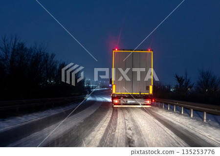 Night winter highway scene freight truck driving icy snow covered road red taillight darkness harsh weather conditions show hard logistics work transport endurance. Cold seasonal industry reality Night winter highway scene freight truck driving icy snow covered road red taillight darkness harsh weather conditions show hard logistics work transport endurance. Cold seasonal industry reality 135253776