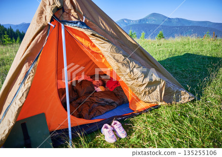 Child sleeping peacefully inside orange and brown tent, snug in sleeping bag on blue mat. Sunlight filters in, illuminating their serene face. Carpathian mountains, Ukraine. 135255106