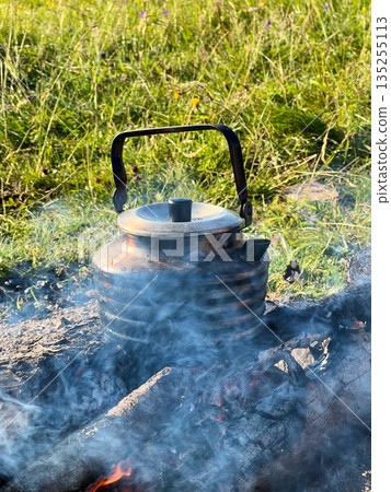 Close-up of metal kettle heating over campfire. Flames and smoke curl around well-used kettle, which rests on logs. Blur of green grass, emphasizing rustic outdoor setting on background. 135255113