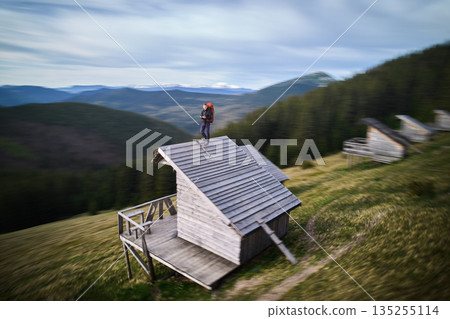 Hiker with orange backpack stands on roof of wooden cabin, surrounded by blurred, dynamic scenery. Motion blur effect. 135255114