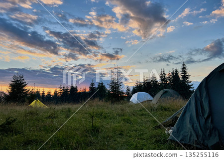 Few tourist tents in grassy field in evening, surrounded by trees and hills under stunning sunset sky. Colorful clouds enhance beauty of scene, with hues of orange and purple illuminating horizon. 135255116