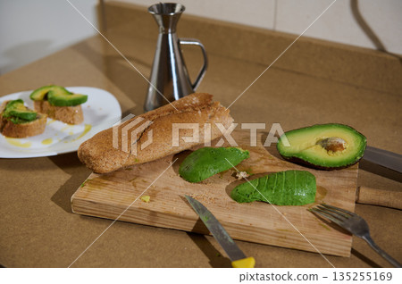 Fresh Avocado Toasts and Crusty Bread on a Wooden Cutting Board with Knife and Oil 135255169