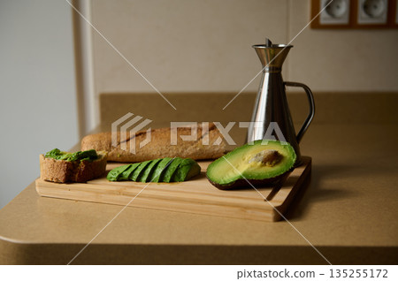Avocado Slices With Whole Grain Bread On Cutting Board And Oil Bottle In Kitchen Scene Avocado Slices With Whole Grain Bread On Cutting Board And Oil Bottle In Kitchen Scene 135255172