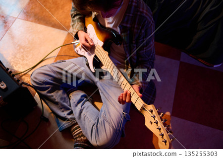 Young Musician Sitting on the Floor Playing Electric Guitar with Amplifier Nearby 135255303