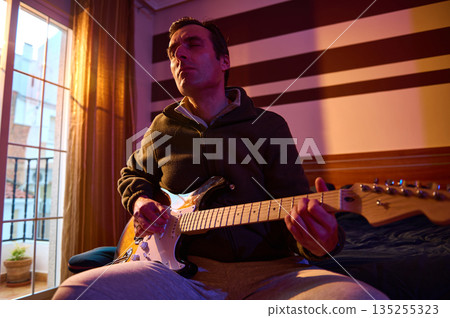 Relaxed Musician Playing Electric Guitar in Cozy Room With Sunset Light and Striped Wall 135255323