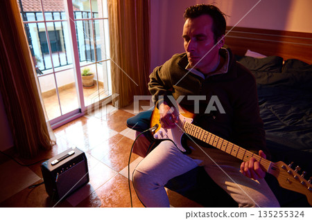 Man Plays Electric Guitar In Cozy Apartment, Focused Practice By Balcony Window With Amplifier Tonight 135255324
