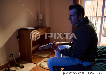 Man Playing Electric Guitar At Home In A Dimly Lit Room Near Window 135255330