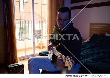 Focused Musician Plays Electric Guitar in a Cozy Bedroom by Balcony Window During Evening Night 135255332