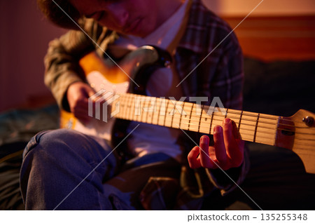 Young Guitarist Plays Electric Guitar In Warm Indoor Setting, Focusing On The Fretboard Young Guitarist Plays Electric Guitar In Warm Indoor Setting, Focusing On The Fretboard 135255348