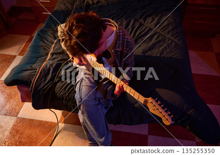 Young Person Strumming Electric Guitar on Bed in Cozy Bedroom with Warm Lighting 135255350
