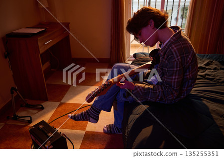 Young Man Playing Guitar While Sitting On Bed In Cozy Bedroom 135255351
