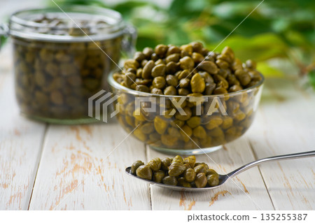 tasty pickled caper buds in a clear glass bowl on a light kitchen table, selective focus. Caper berry in a bowl . tasty pickled caper buds in a clear glass bowl on a light kitchen table, selective focus. Caper berry in a bowl . 135255387