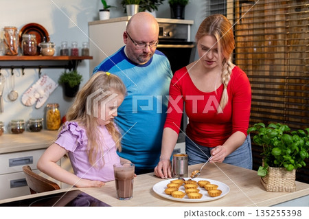 Home kitchen scene with parents and child presenting baked goods, Warm family moment with proud parents and excited child presenting delicious baked treats on white plate 135255398