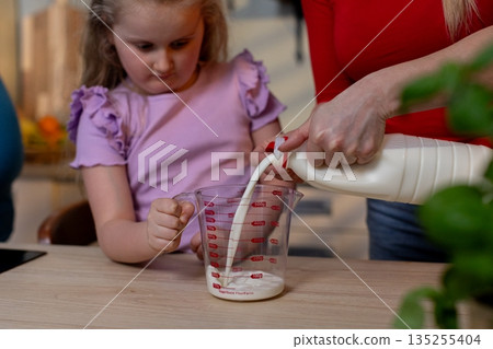 Focused girl transfers milk carefully, Young girl precisely pours milk into container, Focused young girl skillfully pours milk into cup with steady adult supervision Focused girl transfers milk carefully, Young girl precisely pours milk into container, Focused young girl skillfully pours milk into cup with steady adult supervision 135255404
