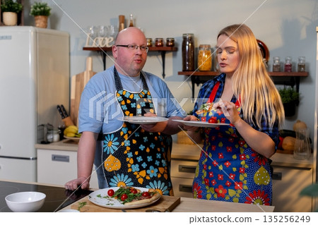 Duo gracefully presents neatly plated dinner in warm setting, Two individuals serve carefully arranged meal with matching aprons and appealing presentation in cozy environment 135256249