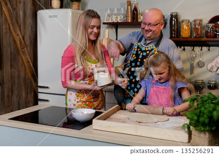 Parents and child tasting batter from mixing bowl, joyful exchange with spoon and bowl, cozy kitchen lighting and decorative jars enhance celebratory home cooking mood 135256291