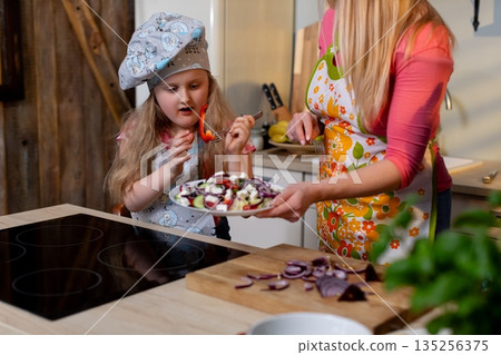 Girl tries dessert with mother, Child explores homemade sweets with guiding parent nearby, Young girl in chef hat enjoying homemade dessert while mother supervises closely Girl tries dessert with mother, Child explores homemade sweets with guiding parent nearby, Young girl in chef hat enjoying homemade dessert while mother supervises closely 135256375
