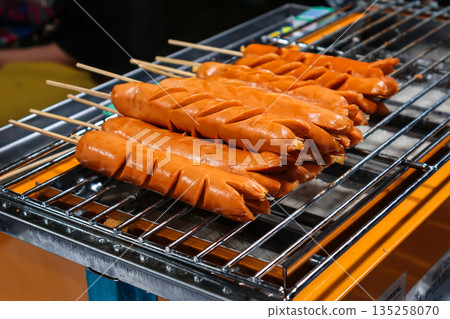 Grilled sausages on skewers with cut marks on a metal rack, street food preparation in natural light for summer picnic Grilled sausages on skewers with cut marks on a metal rack, street food preparation in natural light for summer picnic 135258070