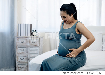 Pregnant Asian woman sitting on bed and gently touching her belly with love and care, representing motherhood, prenatal bonding, and peaceful pregnancy lifestyle in a calm home 135258166