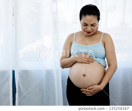 Asian pregnant woman standing indoors, gently holding her bare belly and smiling with love, representing prenatal bonding, maternal care, healthy pregnancy, and emotional connection to unborn baby 135258167