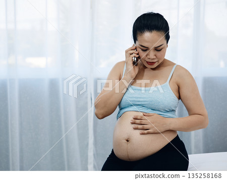Pregnant Asian woman holding her bare belly while talking on the phone, showing concern and care, representing prenatal communication, motherhood, health check, and emotional connection during Pregnant Asian woman holding her bare belly while talking on the phone, showing concern and care, representing prenatal communication, motherhood, health check, and emotional connection during 135258168